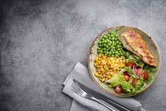 Baked chicken breast on plate with fresh salad, green peas and corn, grey stone background, top