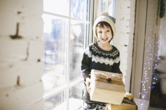 Child sitting at the window on a sunny Christmas day, making presents wrapped in boxes in paper.
