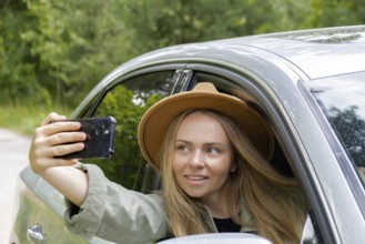 Exited smiling woman making video call with mobile phone from car window. Local solo travel on