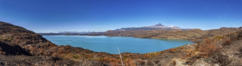 Panoramic photo of a turquoise-coloured lake with surrounding mountains under a blue sky, The