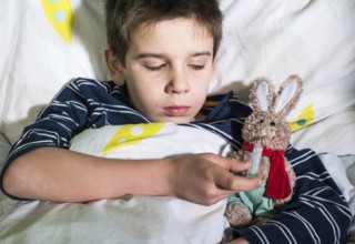 Sick child in bed with teddy bear. Measuring the temperature with a thermometer