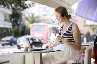Laughing attractive blond woman wearing a headset sitting with a laptop at an open-air restaurant