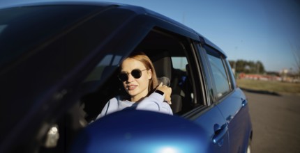 A woman wearing sunglasses drives a blue car on a sunny day