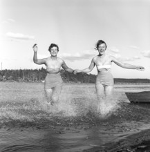 1950s summer. Two young women runs on the shallow part of the beach and the water splashes. Sweden