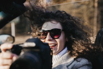 A woman in sunglasses with curly hair smiling holding a camera on a bright day