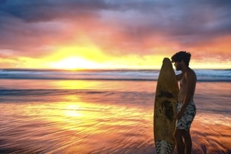 A surfer stands with surfboard in hand on the beach and watches the sun rise over the ocean on a