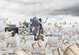 Man in casual wear keeping hand with book up while standing among flying books with cityscape on