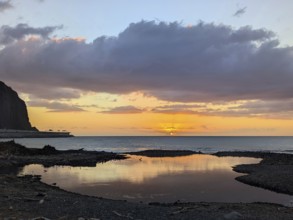 Sunset over the ocean, with striking reflections on wet sand and dramatic clouds in the sky.