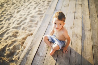 Little Caucasian boy child sitting on wooden pier sandy beach, summer time, sea holiday near water.