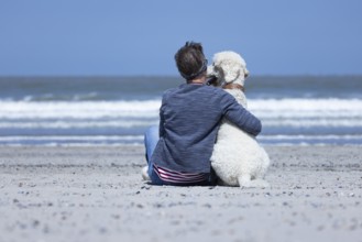 Woman sitting together with her dog, a white poodle, on the beach in the sunshine, hugging him and