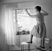 1960s home decorating. A woman at home putting up the curtains in the room window. All by herself