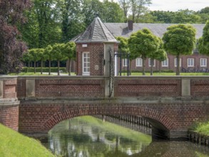 A brick bridge over a body of water, surrounded by manicured hedges and a castle complex,