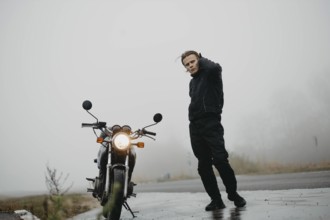 Person standing beside a motorcycle on a foggy road, appearing contemplative
