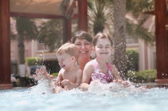 Overjoyed mother with little daughter and son laughing while playing together in swimming pool with