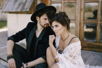 Groom in a black hat sits near bride in a wicker dress on the doorstep of a wooden house.