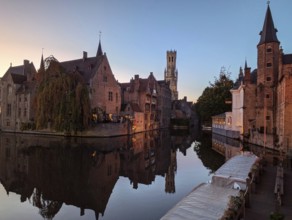 Charming canal at sunset with medieval buildings and a distant tower reflected in the calm water.
