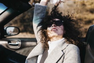 Woman with curly hair enjoys a sunny drive in a convertible, wearing sunglasses and a gray coat