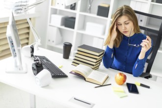 A young girl is sitting at a computer desk, holding glasses in her hand and rubbing her eyes.