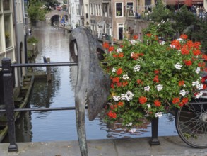 Sculpture and bicycle on a railing along a canal with blooming flowers and reflecting buildings,