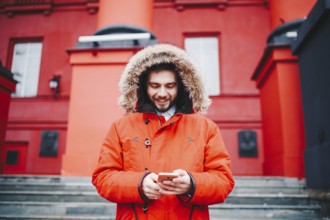 Handsome young male student with toothy smile and beard stands on red wall background, facade of