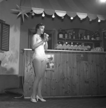Woman in the 1950s. A young woman is standing in a bar with a drink in her hand sipping it with a