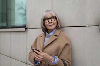 Eine reife Frau in einem hellbraunen Mantel steht an einem modernen Gebäude und überprüft ihr