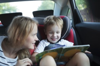 Little boy in a car sitting in child safety seat looking at the pictures in the book, mother