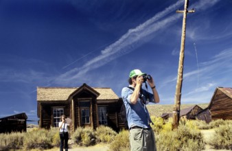 Two tourists photographing and marvelling in front of a wooden house in Bodie. Colour photo,