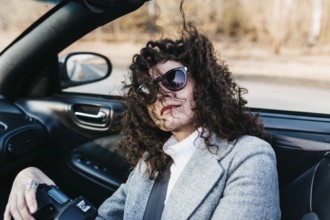 Candid shot of a woman with a camera in a convertible, wind in her curly hair, wearing sunglasses