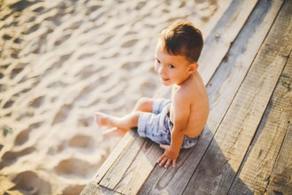 Little Caucasian boy child sitting on wooden pier sandy beach, summer time, sea holiday near water.