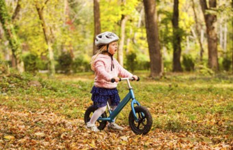 Side view preschool girl in protective helmet riding blue balance bike at the autumn park. Lovely