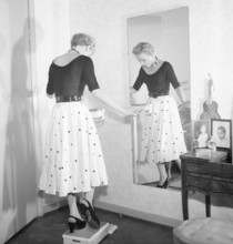 1950s fashion. A young woman is standing on one leg on the scale trying to be as light as possible.