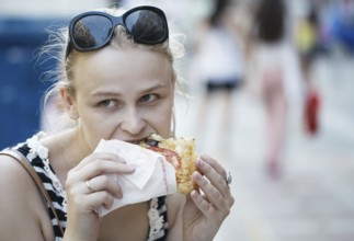 Young woman with sunglasses on her head eating fast food in the street and looking around being