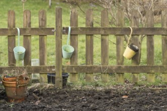 Three empty buckets hanging on a weathered wooden fence in a garden, with a terracotta pot and bare
