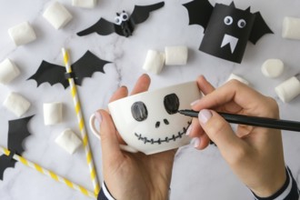 Female hands paint on white cup scary jacks face DIY for kids Halloween home activities Holiday art