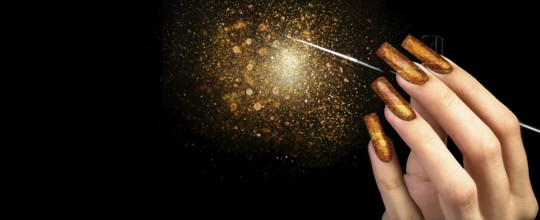 Hand with gold manicure and a brush on a black background covered with sparkles. Nail design.