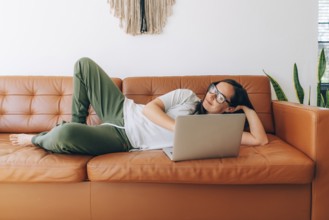 Junge glückliche Frau mit Brille, die zu Hause auf der Couch liegt und an ihrem Laptop arbeitet