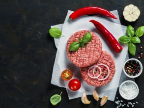 Two raw beef meat steak cutlets for burger with spices and vegetables on black concrete background.