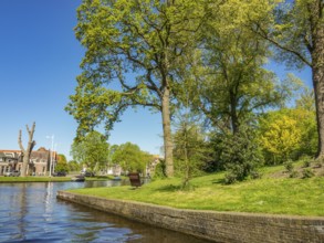 A calm river flows through a park with tall trees and lush green grass under a bright blue sky,