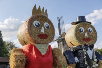Two tall straw men with hats and crowns stand in front of a windmill under a blue sky with clouds,