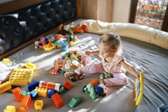 Little girl sits with a xylophone and a wand in her hands on the bed surrounded by soft toys.