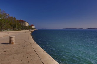 Long promenade along the blue water, with historic buildings and a clear sky in the background,