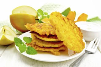 Pumpkin and apple fritters in a white plate, sour cream in a bowl, napkin, mint and fork on wooden