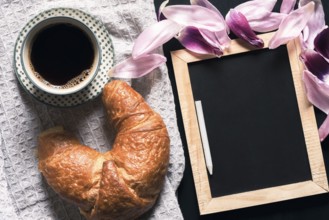Cup of hot coffee and a fresh croissant on a retro kitchen towel, near a blank chalkboard,
