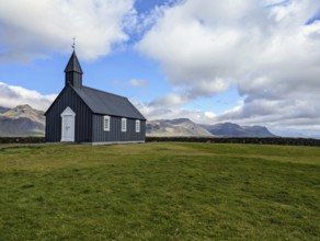 Isolated black church on grassland with mountains in the background under a cloudy sky. Iceland