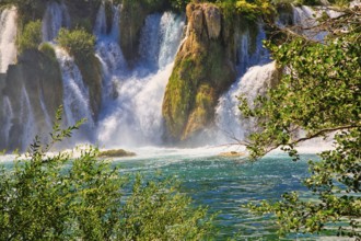 An impressive waterfall flows into a clear, green river, surrounded by lush vegetation and bright