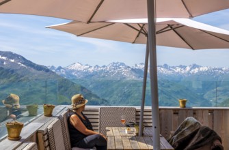 Woman Sitting in a Restaurant Terrace with Mountain View with Snow and Sky in a Sunny Summer Day in