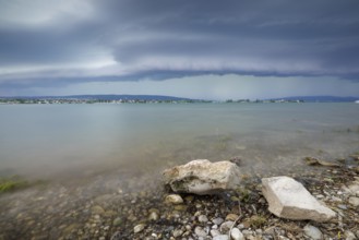Stormy sky over a calm lakeshore with stones and clear water, Allensbach, Untersee, Lake Constance,