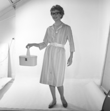 In the 1950s. A woman is pictured in the photographers studio wearing a dress and holding a handbag