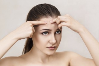 Young girl squeezes pimple on her forehead over beige background